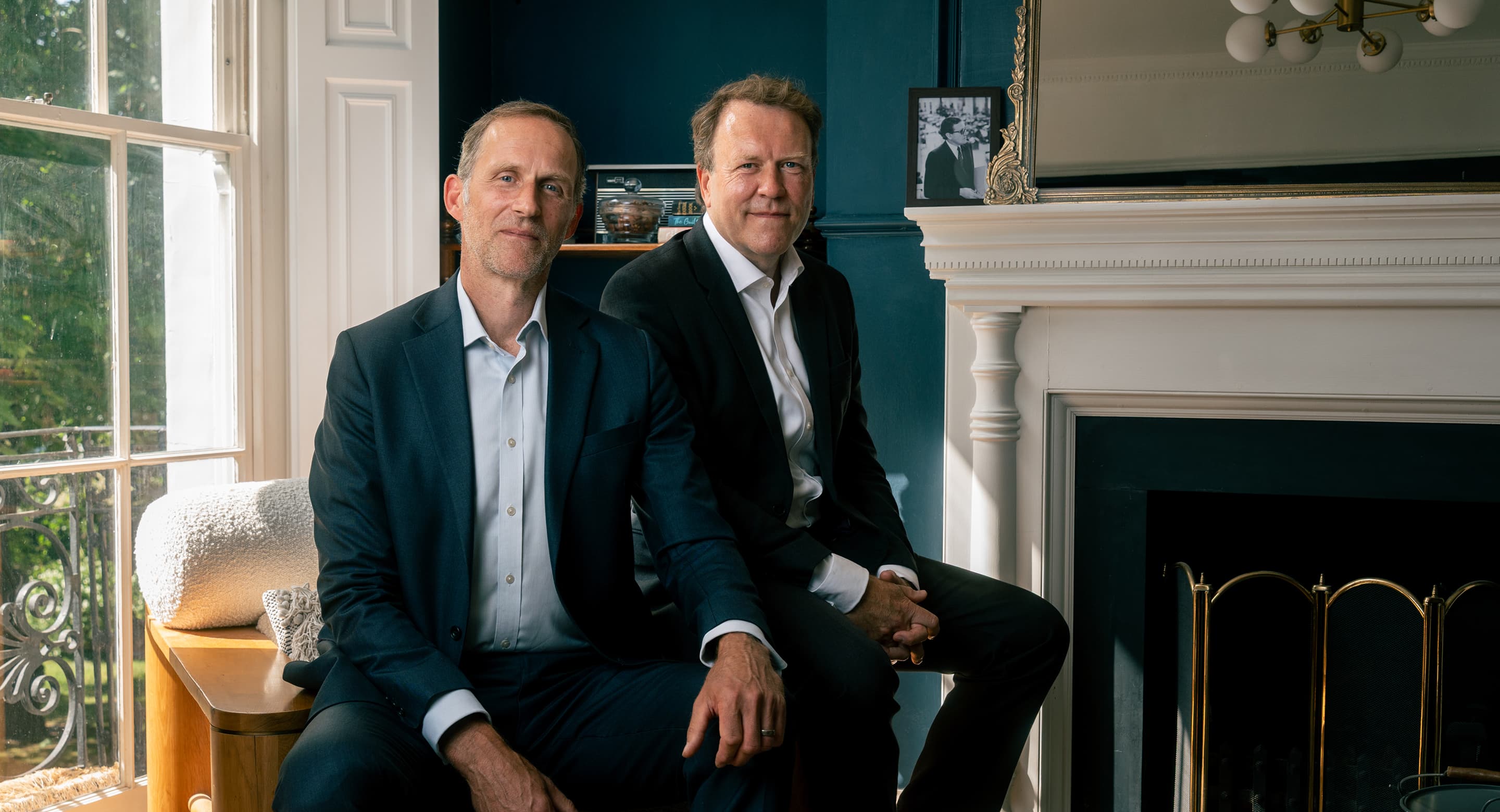 Two CJ Coleman directors seated in a bright office with a classic fireplace and large window.
